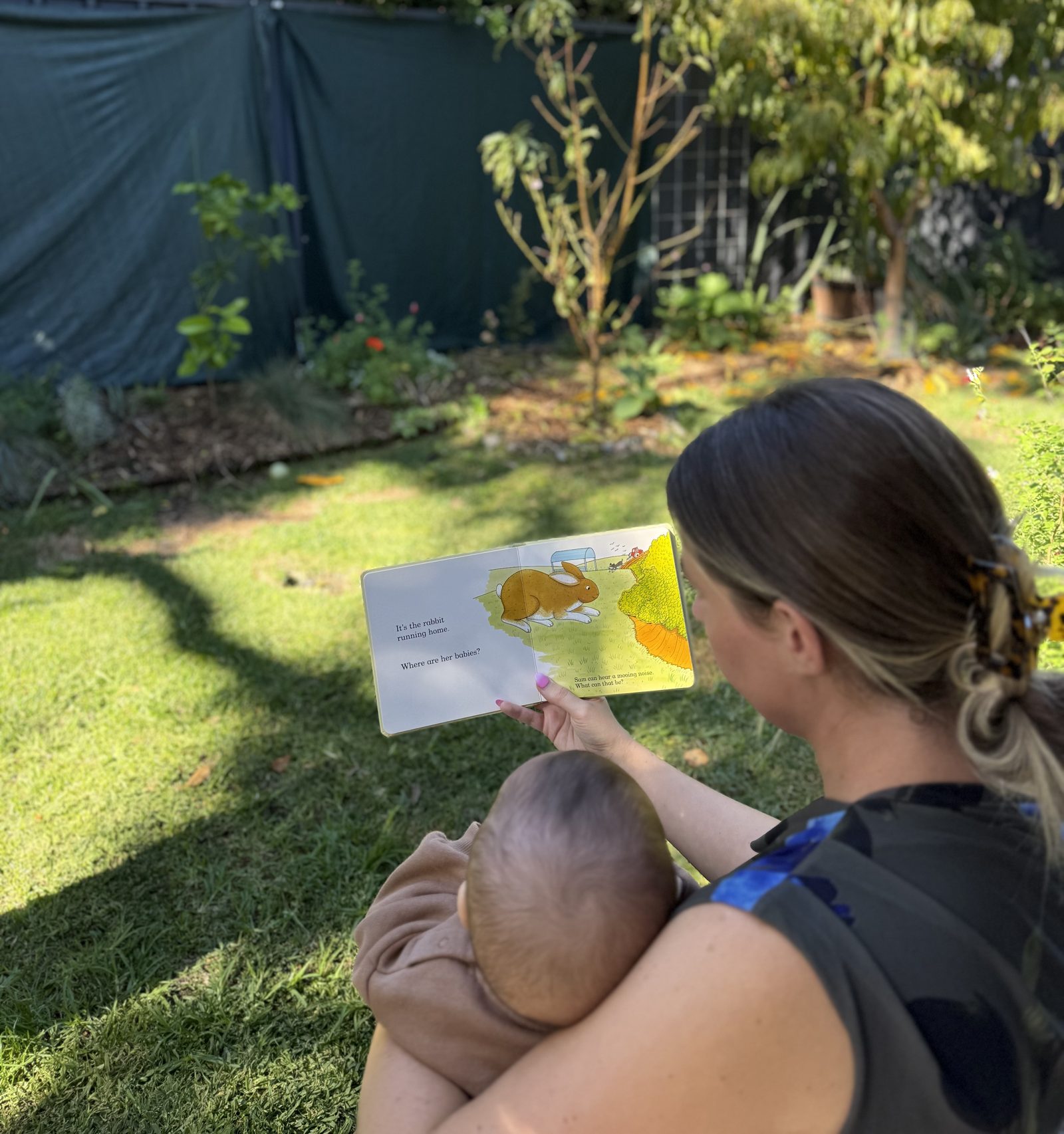 Speech pathologist seated on grass reading a board book to a baby on her lap
